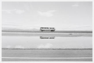 A greyhound bus travels across a landscape and is reflected in a body of water.