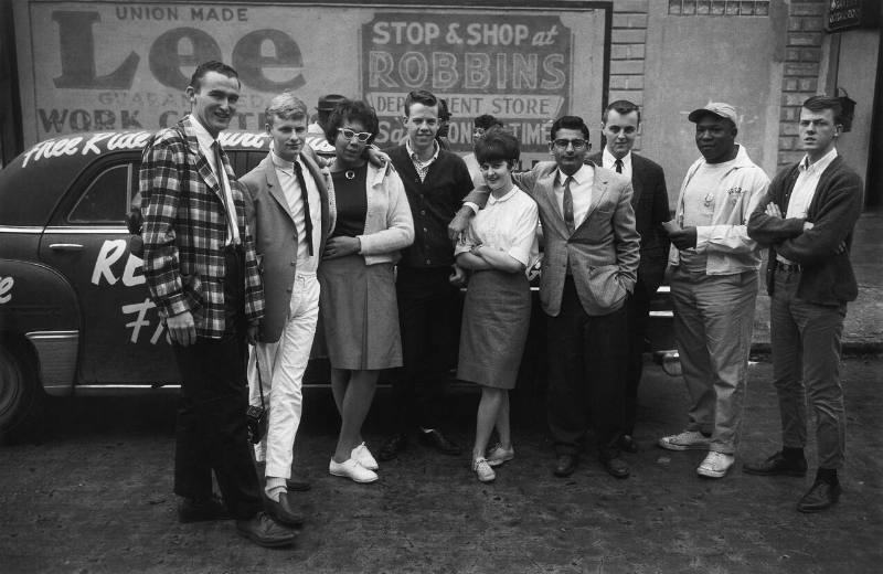 A black and white photograph of a group of individuals standing in front of a car painted with …
