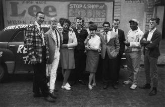 A black and white photograph of a group of individuals standing in front of a car painted with …