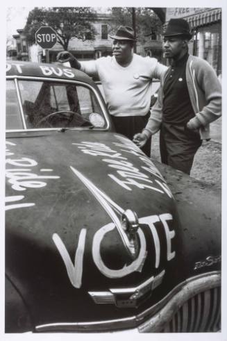 A black and white photograph of two men leaning against the side of a car with "BALLOT BUS" wri…
