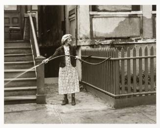 A woman standing between a staircase and a fenced yard on a sidewalk sprays water from a hose w…