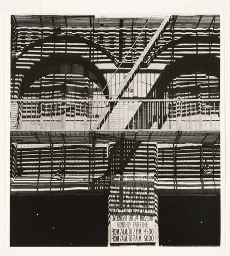 A black and white photograph shows a heavily-shadowed building with a fire escape in the center…