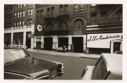 A black and white photograph showing two cars parked on a street across from a store front with…