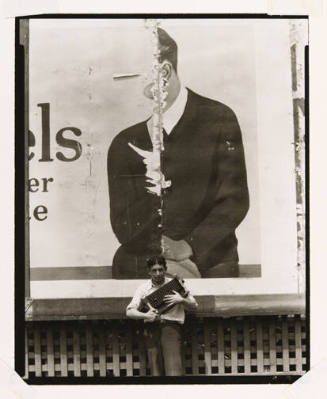 A black and white photograph of a young man holding a camera in front of a ragged billboard.
