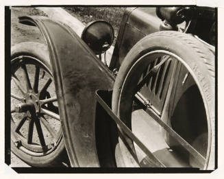 A black and white image of the front side of a car and the curb of a nearby sidewalk.