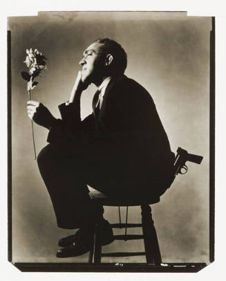 A black and white photograph of a man sitting on a stool looking at a flower and a gun sticking…