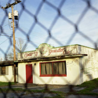 A color photograph of an old Shriner's Club building of white brick with red window and door as…