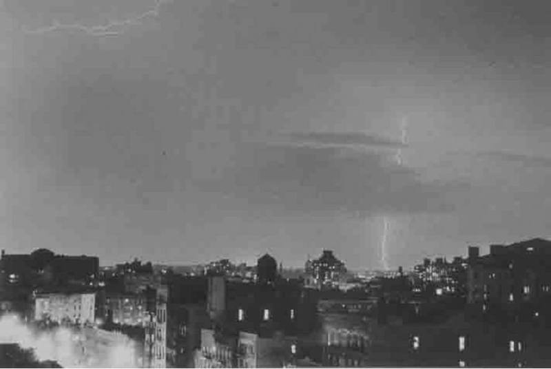 A black and white photograph of a nighttime, rooftop view of buildings and two lightning strike…