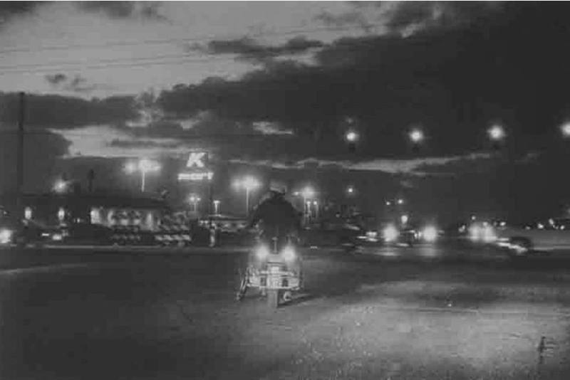A black and white photograph of a street intersection at night. A police officer on a motorcycl…