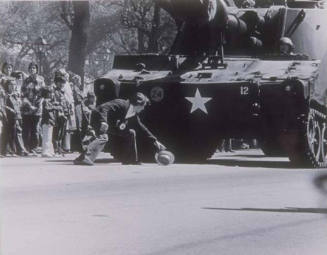 A photograph of a man retrieving his hat from in front of a military tank.