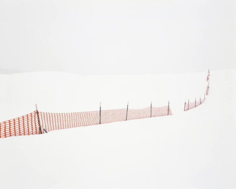 A color photograph of a snow-covered landscape interrupted by an orange plastic fence.

