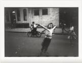 A black and white photograph of an urban scene with a young boy standing with his arms stretche…