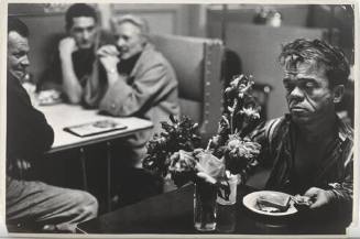 A black and white photograph of Jimmy Armstrong eating a sandwich at a diner with a vase of flo…