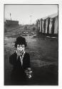 A black and white photograph of a little person wearing clown make-up and smoking outside of a …