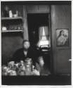 A black and white photograph of a woman seated at a table covered in kitchenwares in front of a…