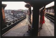 A color photograph of two women standing and reading in brightly colored dresses while waiting …