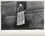 A black and white photograph of a woman leaning out of her window with a flower pot to her righ…