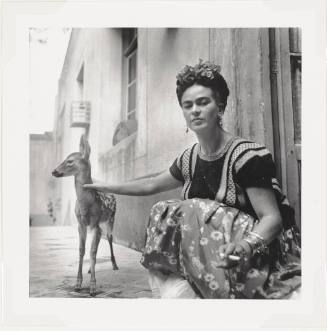 A black and white photograph of Frida Kahlo squatting next to a building with one hand on the n…