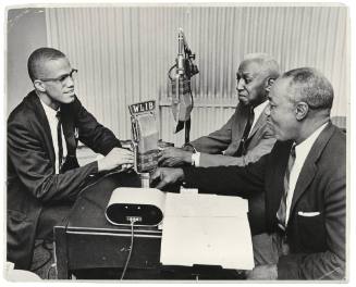 A black and white photograph of three men seated around a table with microphones conversing.