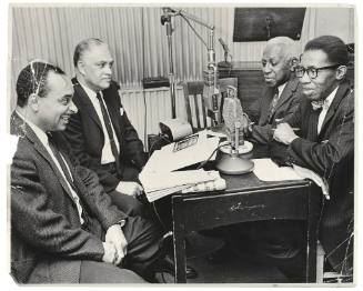 A black and white photograph of four middle-aged men sitting around a table with two microphone…