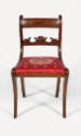 A frontal view of a Grecian-style klismos side chair with a red cushion decorated in a gold fol…