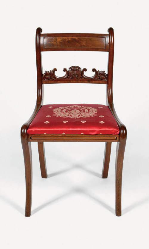 A frontal view of a Grecian-style klismos side chair with a red cushion decorated in a gold fol…