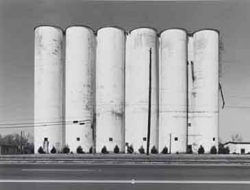 A black and white photograph of a series of white grain silos.

