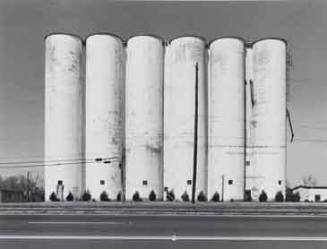 A black and white photograph of a series of white grain silos.

