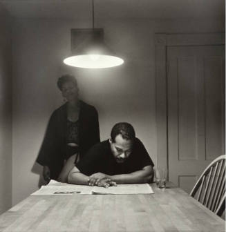 A black and white photograph of a man sitting at a wooden table reading the newspaper under the…