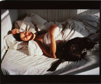 A color photograph of a sleeping woman wrapped in bed sheets on a sun-dappled bed with a black …