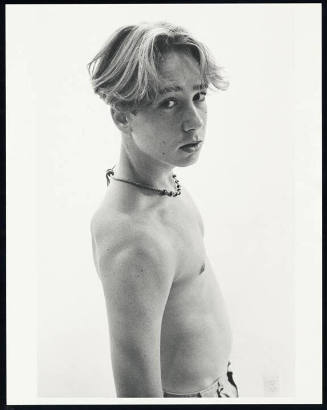 A black and white three-quarters length portrait of a shirtless young man standing in partial p…