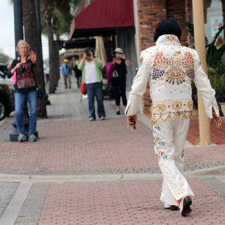 A man walking down the street dressed as the late Elvis Presley in a white, bedazzled suit. 