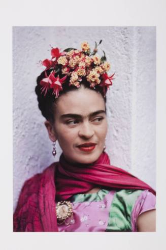 A color photograph showing Frida Kahlo posed against a white wall and dressed in bright pink an…