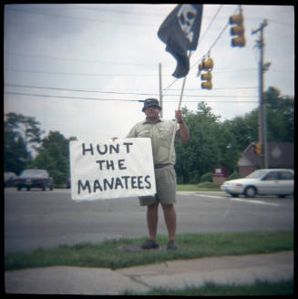 A man standing next to a road holding a pirate's flag and a sign with the phrase “Hunt the Mana…
