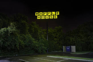 A large, backlit yellow Waffle House sign at the edge of an empty parking lot by a blue dumpste…