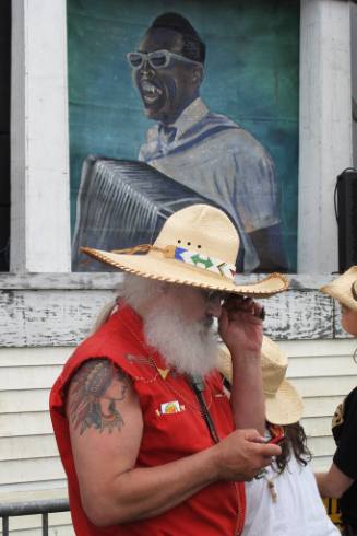 A man with a bushy white beard wearing a bright red sleeveless shirt and a wide-brimmed straw h…