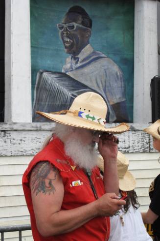 A man with a bushy white beard wearing a bright red sleeveless shirt and a wide-brimmed straw h…