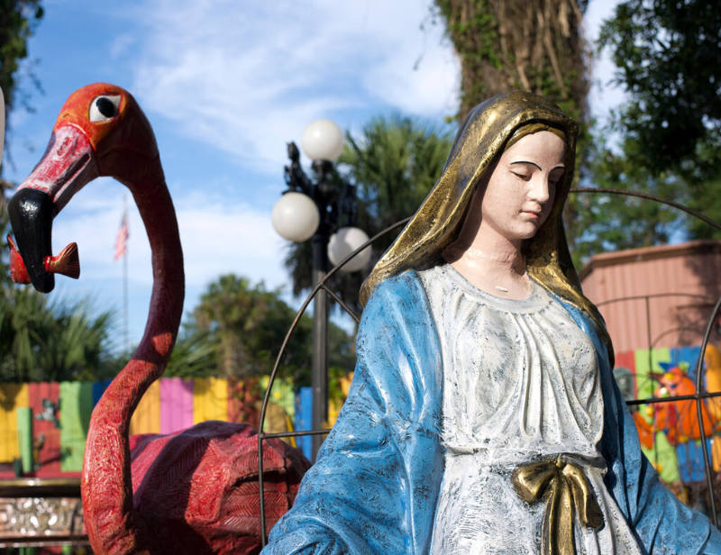 A flamingo sculpture adjacent to a Virgin Mary sculpture wearing a blue and white robe with a g…