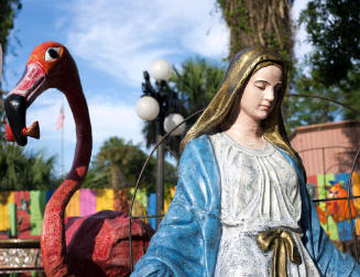 A flamingo sculpture adjacent to a Virgin Mary sculpture wearing a blue and white robe with a g…